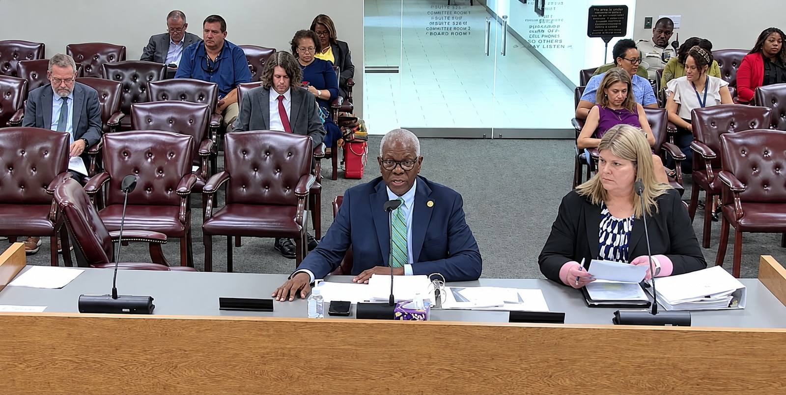 Shelby County Trustee Joseph Lee III speaks into a microphone at a hearing table, with another presenter beside him and attendees seated behind them.