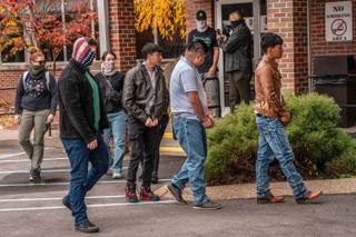 Five people in handcuffs and leg restraints are escorted by masked officers across a parking lot outside a county courthouse.
