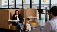An Escape Lounge interior with upholstered booth seating, a traveler with a coffee cup, and floor-to-ceiling windows overlooking an airport apron.