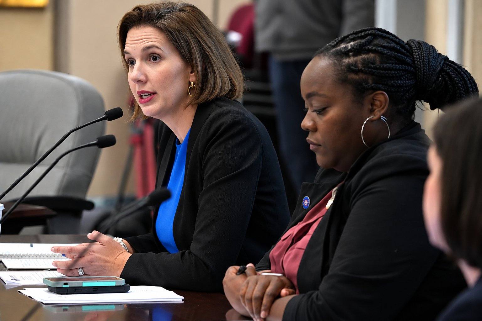 Women seated at a formal hearing table, with one speaking into microphones and another taking notes.