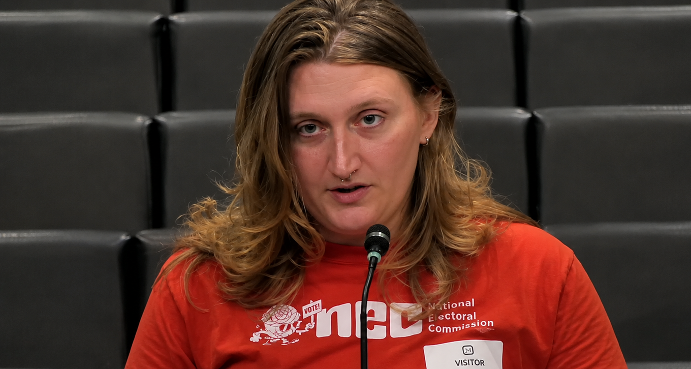 A woman in a red National Electoral Commission t-shirt speaks into a microphone at a Memphis City Council public comment podium.