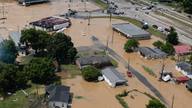 Aerial view of a flooded small Tennessee town, with muddy brown water covering streets, yards, homes and a strip of storefronts beside a highway.