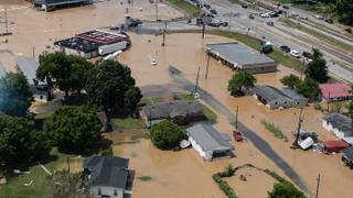 Aerial view of a flooded small Tennessee town, with muddy brown water covering streets, yards, homes and a strip of storefronts beside a highway.