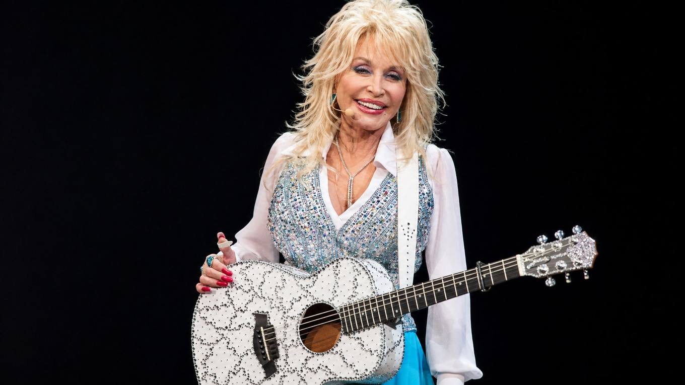 Dolly Parton on stage holding a white rhinestone acoustic guitar.