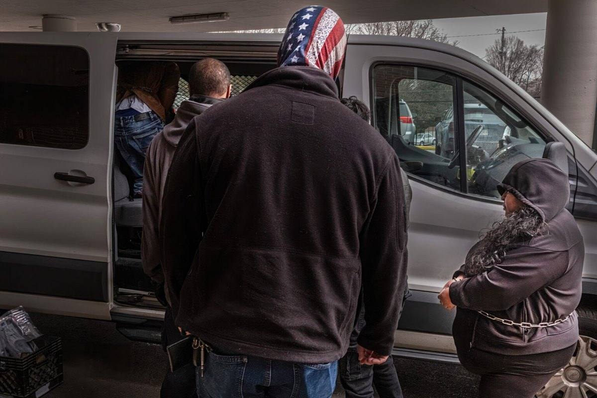 Masked agents stand beside a white transport van as detained people, one in handcuffs, are loaded into it outside a courthouse.