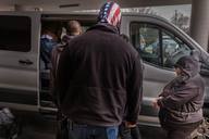 Masked agents stand beside a white transport van as detained people, one in handcuffs, are loaded into it outside a courthouse.
