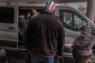Masked agents stand beside a white transport van as detained people, one in handcuffs, are loaded into it outside a courthouse.