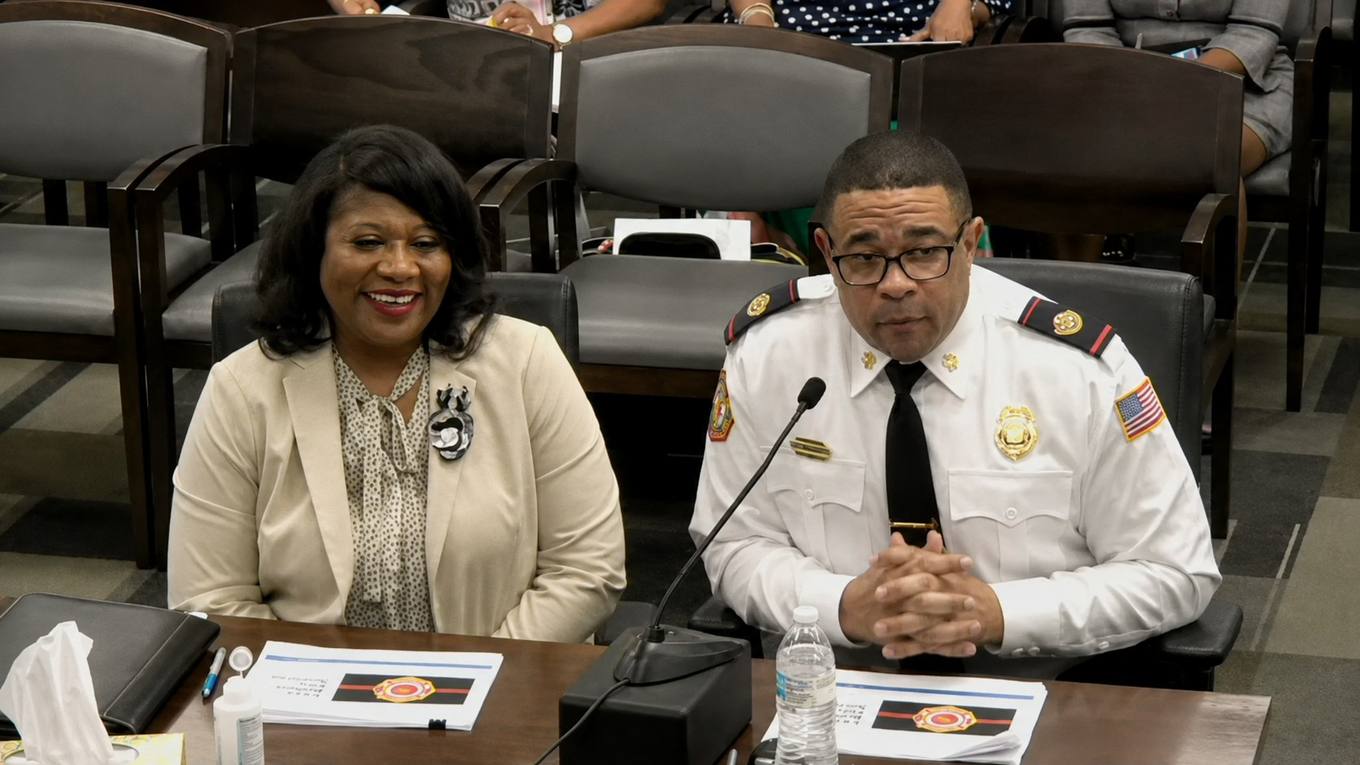 Two city officials seated at a witness table during a Memphis City Council budget hearing, one wearing a fire department uniform.