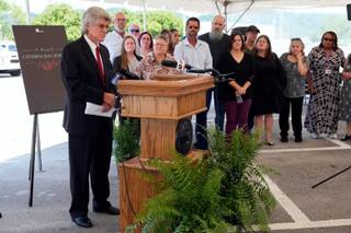 Dennis Jenkins speaks at a podium with news microphones outside Riverbend Maximum Security Institution as family members stand behind him near a memorial sign.