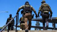 Officers wearing Police ICE vests stand and climb stairs beside a parking garage under a clear blue sky.
