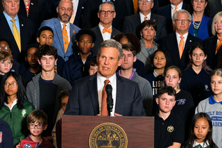 Gov. Bill Lee speaks at a podium bearing the Tennessee state seal, flanked by students and Republican lawmakers wearing orange ties, as he signs the Memphis-Shelby County Schools takeover bill into law.