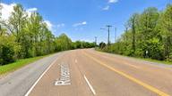 A curving roadway labeled Riverport Road, lined with trees and utility poles under a blue sky.