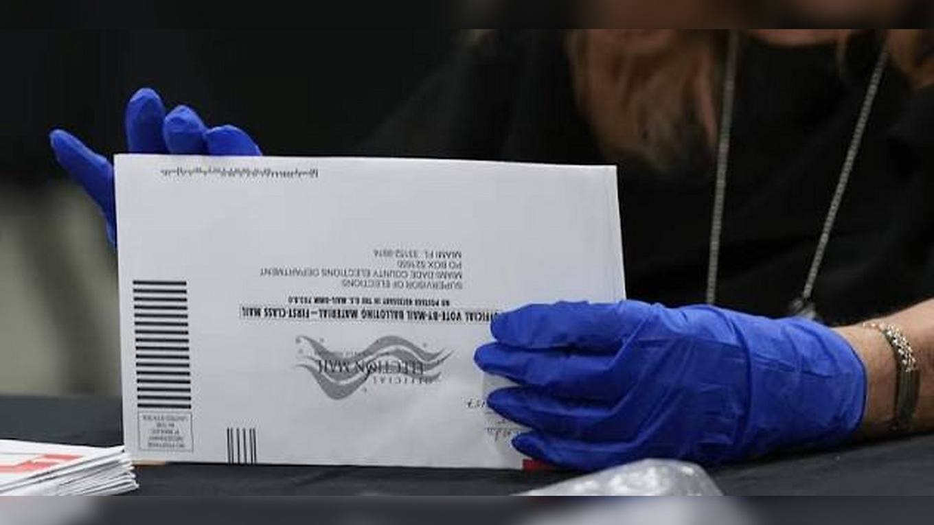 A gloved election worker handles a white official vote-by-mail ballot envelope at a counting table.