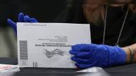 A gloved election worker handles a white official vote-by-mail ballot envelope at a counting table.
