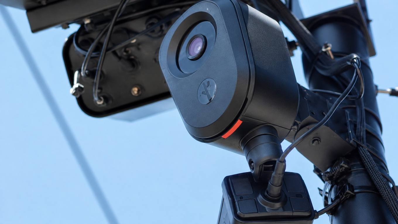 A small black camera on a slim roadside pole above a blurred stream of passing traffic under an overcast sky.