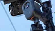 A small black camera on a slim roadside pole above a blurred stream of passing traffic under an overcast sky.