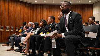City officials and uniformed police leadership seated in a wood-paneled council chamber gallery, waiting to be heard.