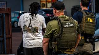 Federal agents in U.S. Marshal and FBI vests stand with a handcuffed man during a street arrest.