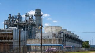 A row of industrial gas turbine generator units behind chain link fencing under a hazy sky, representing the temporary turbines scheduled for removal.