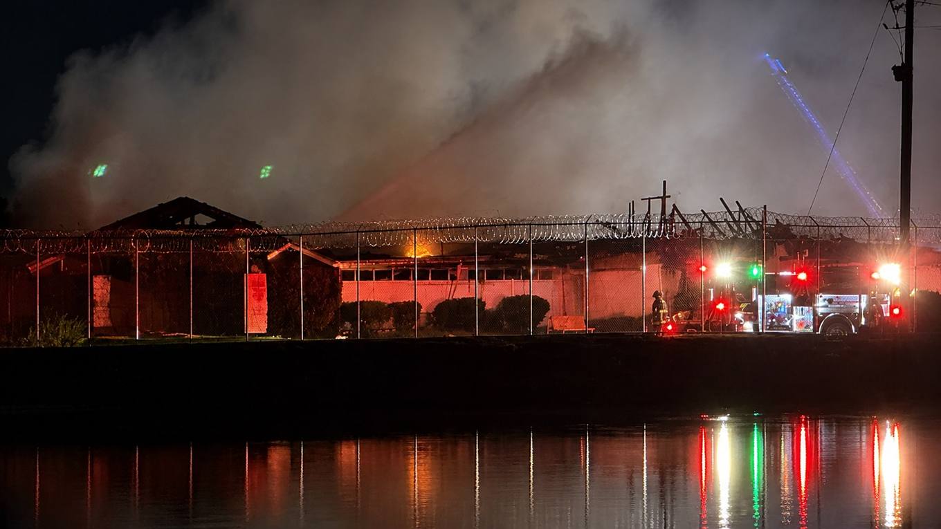 Emergency crews and a fire engine at a burning low institutional building behind a razor wire fence at night.
