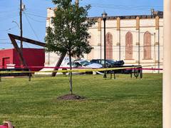 Wrecked vehicles behind yellow and red caution tape beside a grassy area, a tree, and buildings in daylight.