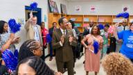 Officials, educators and supporters clap and wave blue and red pom-poms during a celebration in a school library with bookshelves behind them.