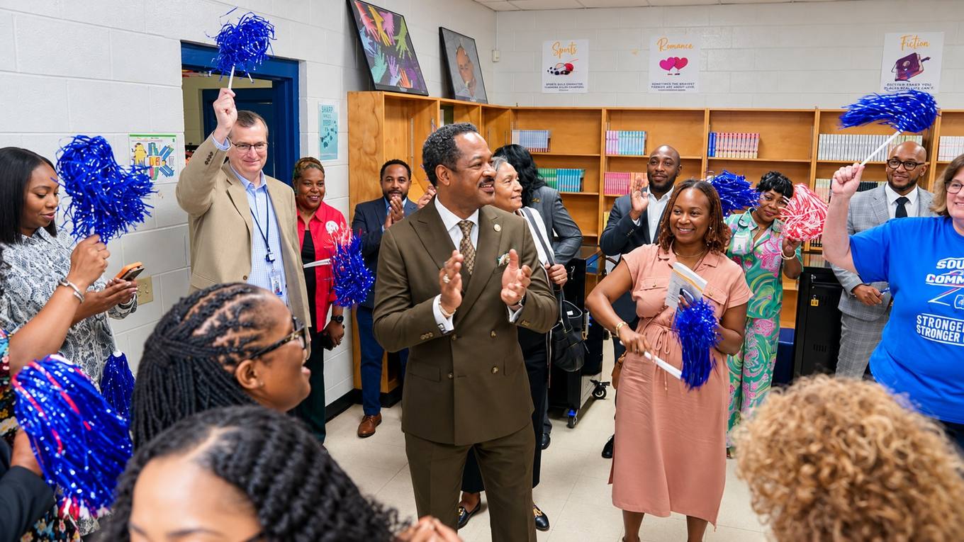 Officials, educators and supporters clap and wave blue and red pom-poms during a celebration in a school library with bookshelves behind them.