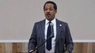 A man in a pinstriped suit speaks into a microphone at a wooden podium during a public meeting.