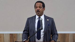 A man in a pinstriped suit speaks into a microphone at a wooden podium during a public meeting.