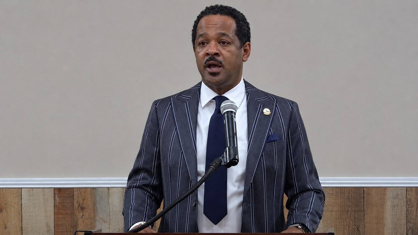 A man in a pinstriped suit speaks into a microphone at a wooden podium during a public meeting.