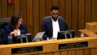 A county commissioner at the dais and a second commissioner reviewing a document during a commission meeting.