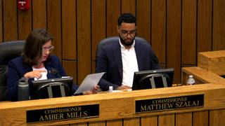 A county commissioner at the dais and a second commissioner reviewing a document during a commission meeting.