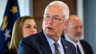 A man in a dark suit and glasses speaks at a public briefing with officials seated behind him and a flag in the background.