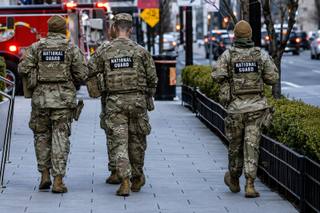 Three National Guard members in camouflage uniforms walking along a city sidewalk past emergency vehicles.
