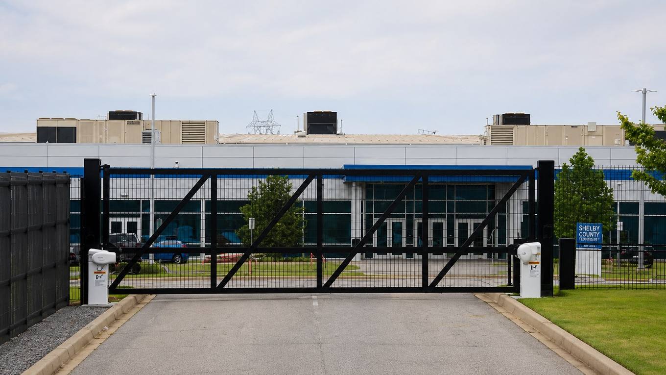 A closed black security gate across a driveway in front of a large data center building with rooftop cooling units.