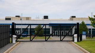 A closed black security gate across a driveway in front of a large data center building with rooftop cooling units.