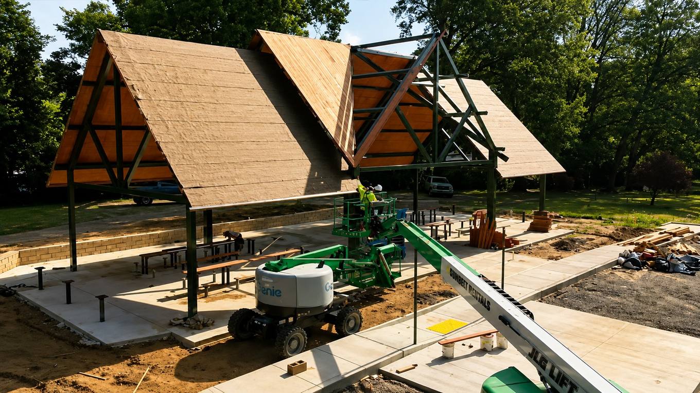 A nearly finished public park pavilion under construction, with new steel framing and roof decking, a lift on site and fresh concrete walkways.