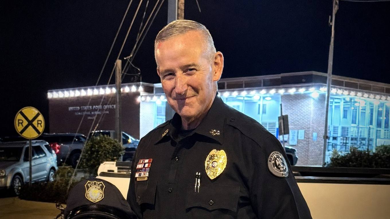 A uniformed police chief in a dark uniform with a gold badge stands at night outside a lighted building, holding his cap.