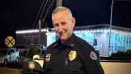 A uniformed police chief in a dark uniform with a gold badge stands at night outside a lighted building, holding his cap.