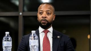 A county official in a dark suit and red tie sits at a table with water bottles and a microphone during a public meeting.