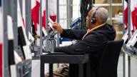 An election official wearing headphones uses a stylus to inspect a voting machine among rows of election equipment.
