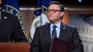 House Speaker Mike Johnson stands at a U.S. Capitol news conference holding a leather folder, with U.S. House flags behind him.