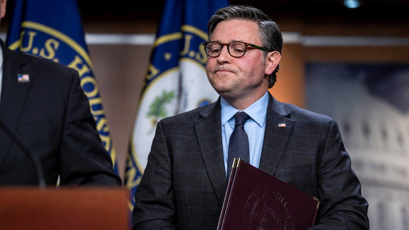 House Speaker Mike Johnson stands at a U.S. Capitol news conference holding a leather folder, with U.S. House flags behind him.