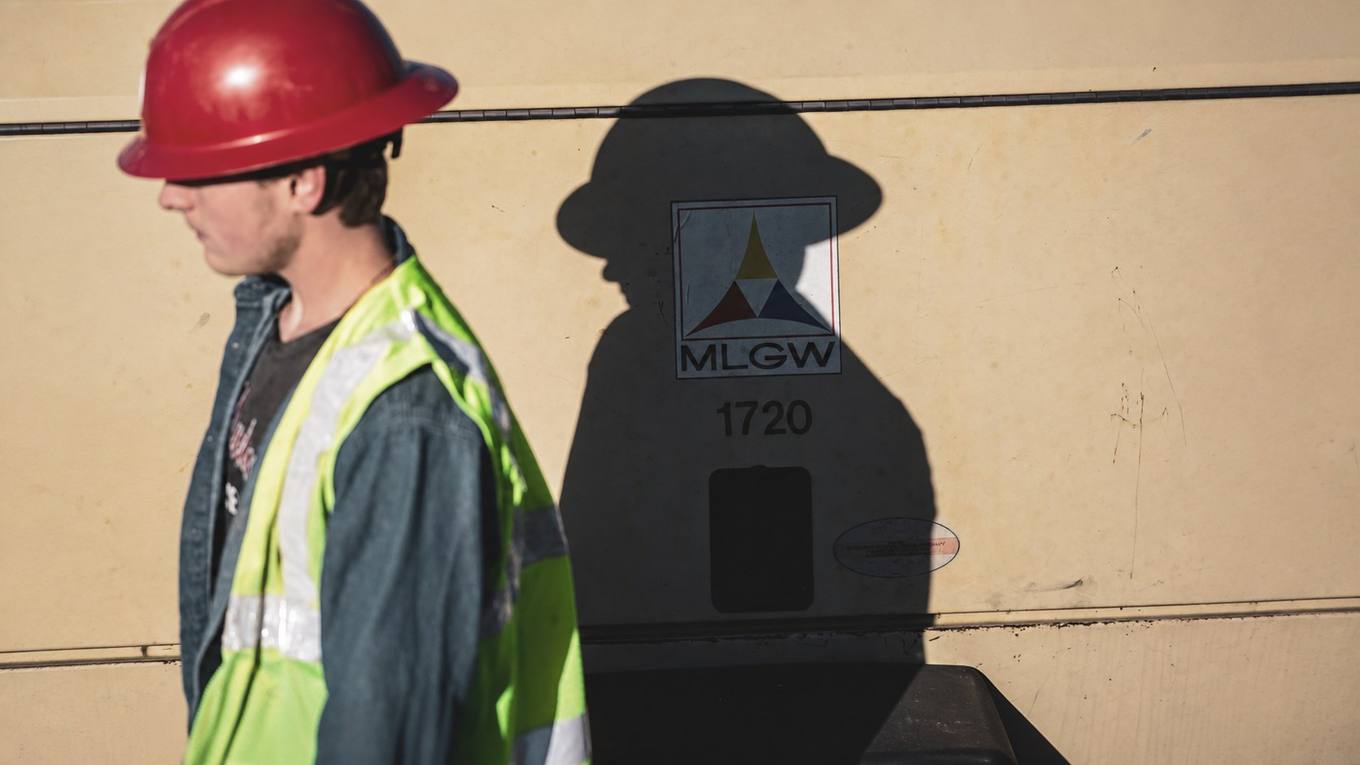Memphis Light, Gas and Water worker in a red hard hat and reflective vest beside an MLGW truck during storm restoration work.