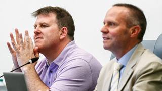Two men seated at a public meeting dais, one resting his hands near a microphone while the other speaks.