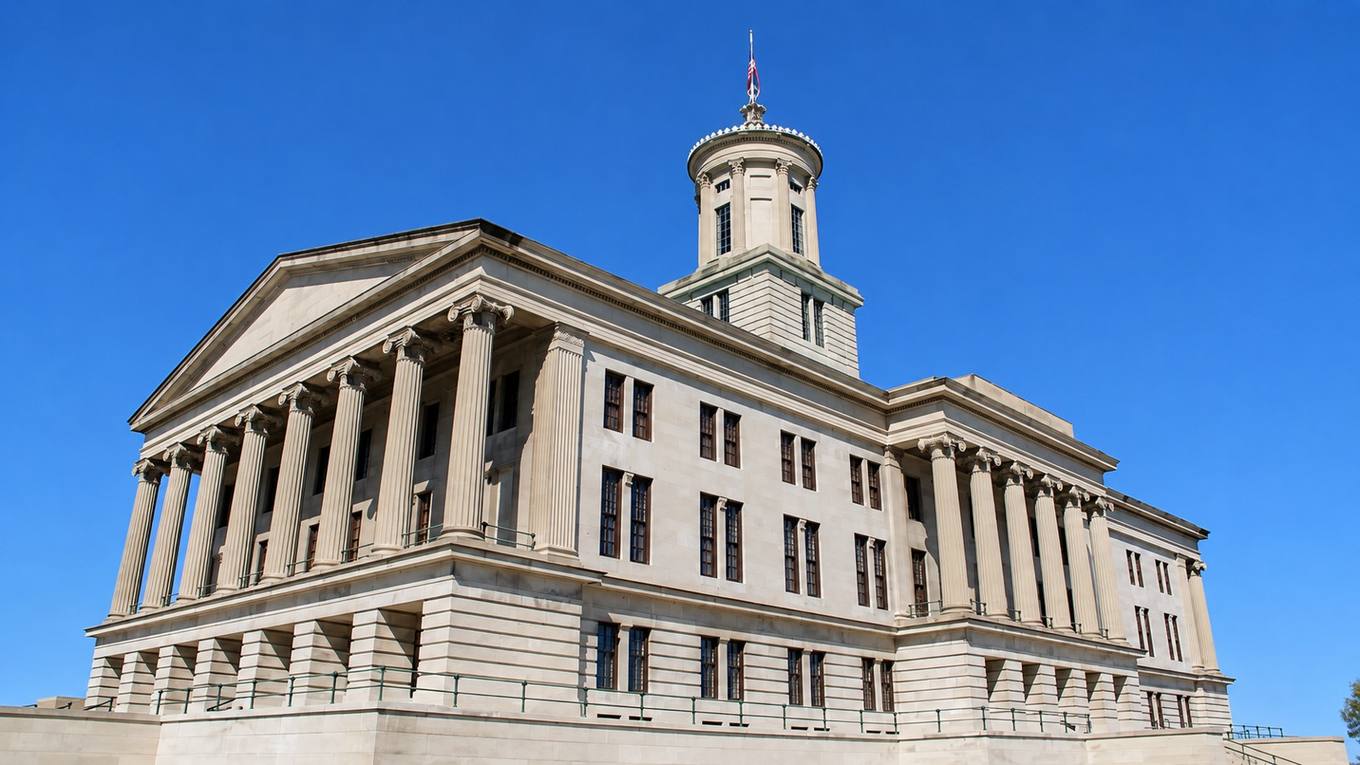 The Tennessee State Capitol in Nashville, with its columned facade, cupola, and flag under a clear blue sky.