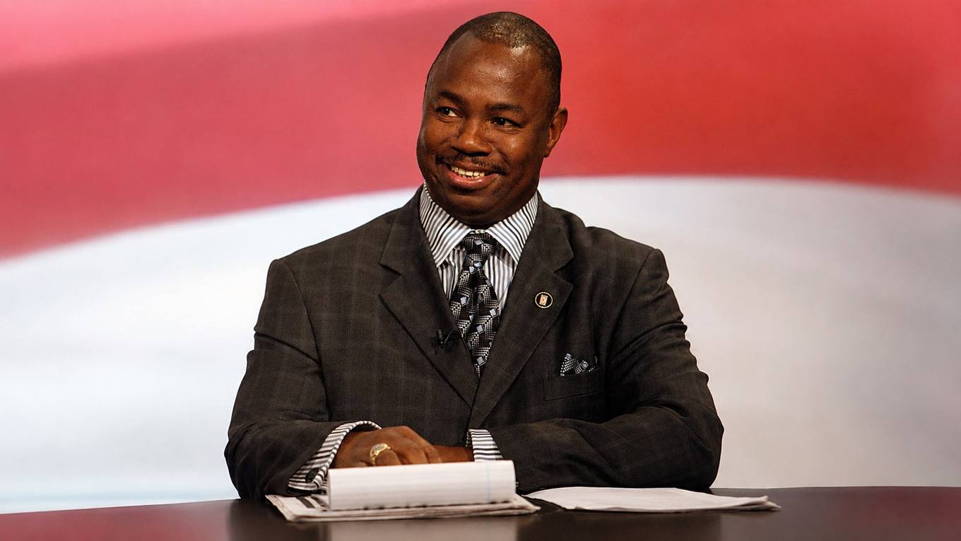 Joe Towns Jr. seated at a table in a suit, smiling, with an American flag backdrop behind him.
