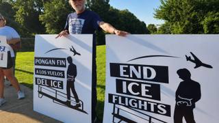 A demonstrator holds two large protest signs reading "End ICE Flights" and "Pongan Fin a los Vuelos del ICE" on a grassy roadside, with another participant standing nearby.